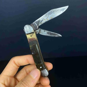 May include: A vintage folding pocket knife with two weathered steel blades and a dark, tarnished metal handle. The handle features a brass-toned bolster near the pivot point and a rounded metallic end cap, showing signs of age and oxidation.