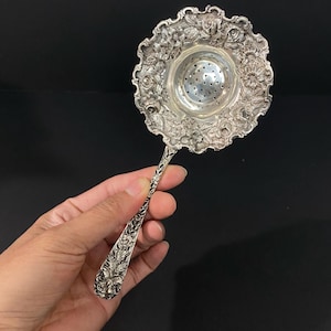 May include: A silver-toned metal tea strainer featuring a circular bowl with a perforated center and a scalloped edge decorated with embossed floral patterns. The handle is long and slender, also adorned with intricate floral and leaf engravings.