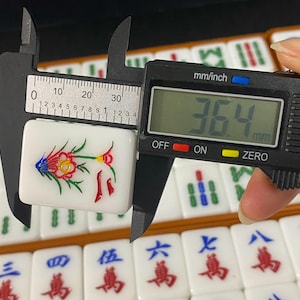 May include: A white Mahjong tile with a colorful floral design is measured with a digital caliper. The tile is approximately 3.6 cm long. Other tiles with Chinese characters are visible in the background.
