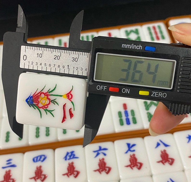 May include: A white Mahjong tile with a colorful floral design is measured with a digital caliper. The tile is approximately 3.6 cm long. Other tiles with Chinese characters are visible in the background.