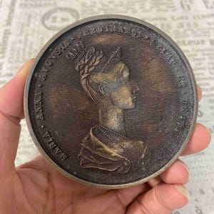May include: A circular bronze-toned commemorative medallion featuring a relief profile of a woman wearing a crown and necklace. The text MARIA ANNA AVGVSTA FERDINANDI I IMP. ET REGIS is embossed along the raised outer edge of the metal disc.