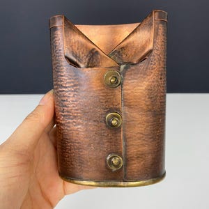 Op de afbeelding: Een koperkleurige, cilindrische container met een getextureerd oppervlak, die op een vest lijkt. Het heeft een kraagachtig bovenstuk en drie messingkleurige knopen aan de voorkant. De basis heeft een messingkleurige rand.