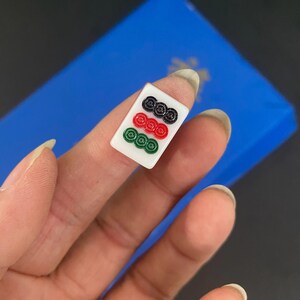 Puede incluir: Una ficha de Mahjong blanca con tres filas de s&iacute;mbolos circulares en negro, rojo y verde. La ficha se sostiene sobre un fondo azul.