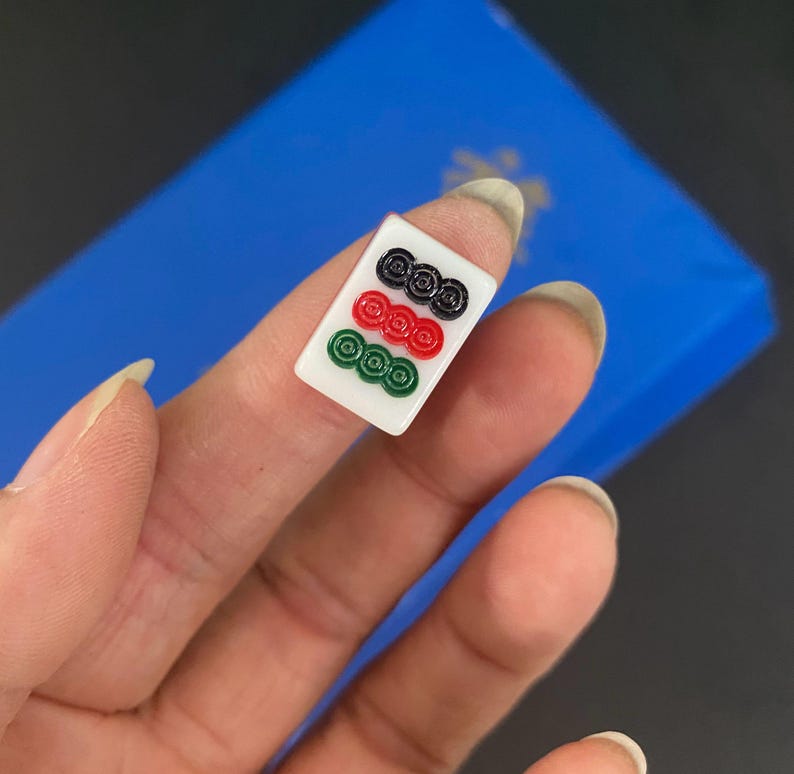 Puede incluir: Una ficha de Mahjong blanca con tres filas de s&iacute;mbolos circulares en negro, rojo y verde. La ficha se sostiene sobre un fondo azul.