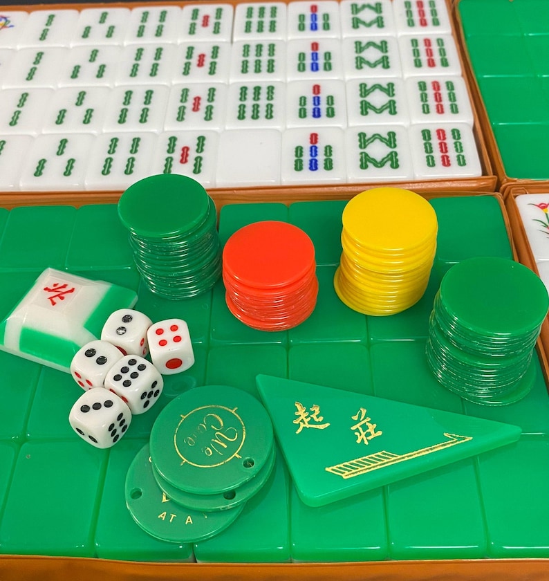 May include: A Mahjong set with white tiles, green and blue symbols, dice, and coloured chips. The set includes green, red, and yellow chips, a green triangular marker and dice.