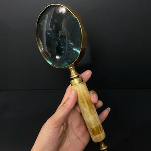 May include: A magnifying glass featuring a circular glass lens encased in a polished brass frame. The handle is constructed from segments of cream and tan marbled material, accented with brass fittings at the top and a small brass finial at the base.