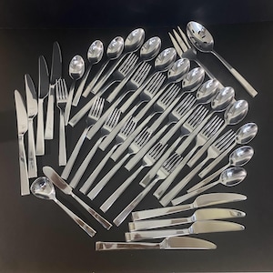 Puede incluir: Un gran conjunto de cubiertos de acero inoxidable pulido que incluye cuchillos de mesa, tenedores, cucharas y una cuchara de servir ranurada. El set presenta un diseño elegante con mangos rectos y superficies reflectantes agrupadas.