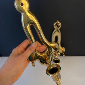 May include: A pair of vintage brass candle holders. One is held by a hand, the other is in the background. The candle holders have a curved design with a circular opening and a cup for the candle. The brass has a shiny finish.