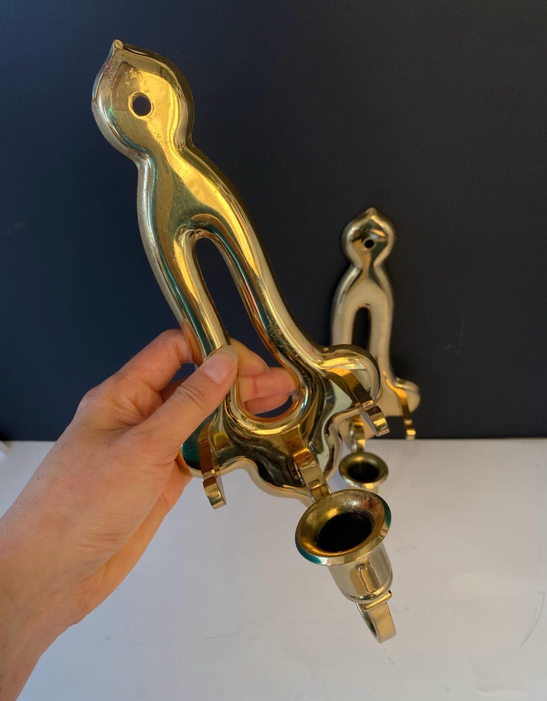 May include: A pair of vintage brass candle holders. One is held by a hand, the other is in the background. The candle holders have a curved design with a circular opening and a cup for the candle. The brass has a shiny finish.