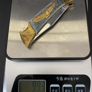 May include: A folding pocket knife with a gold and silver handle. The handle features an engraved eagle design. The knife is resting on a digital scale, which reads 99.3.