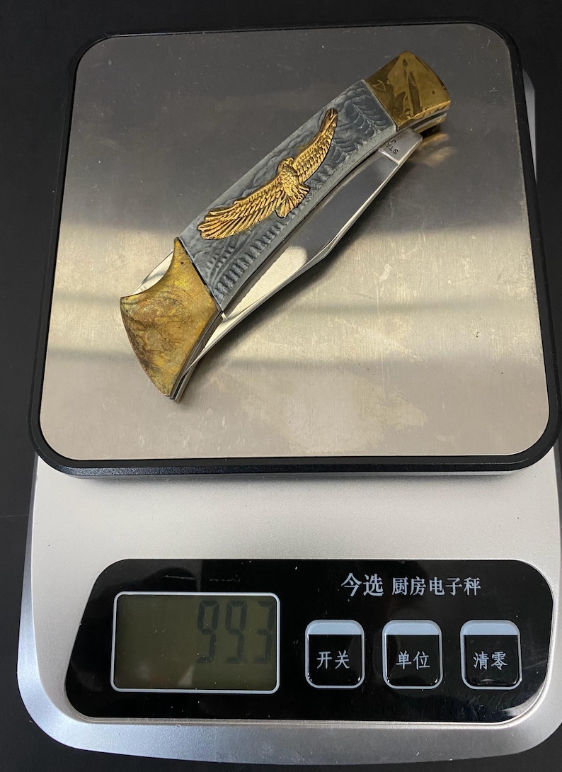 May include: A folding pocket knife with a gold and silver handle. The handle features an engraved eagle design. The knife is resting on a digital scale, which reads 99.3.