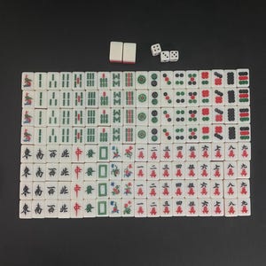Puede incluir: Un juego de Mahjong completo con fichas blancas que presentan varios s&iacute;mbolos y caracteres en rojo, verde y negro. Incluye cuatro dados y dos fichas de repuesto. Las fichas est&aacute;n dispuestas sobre una superficie oscura.
