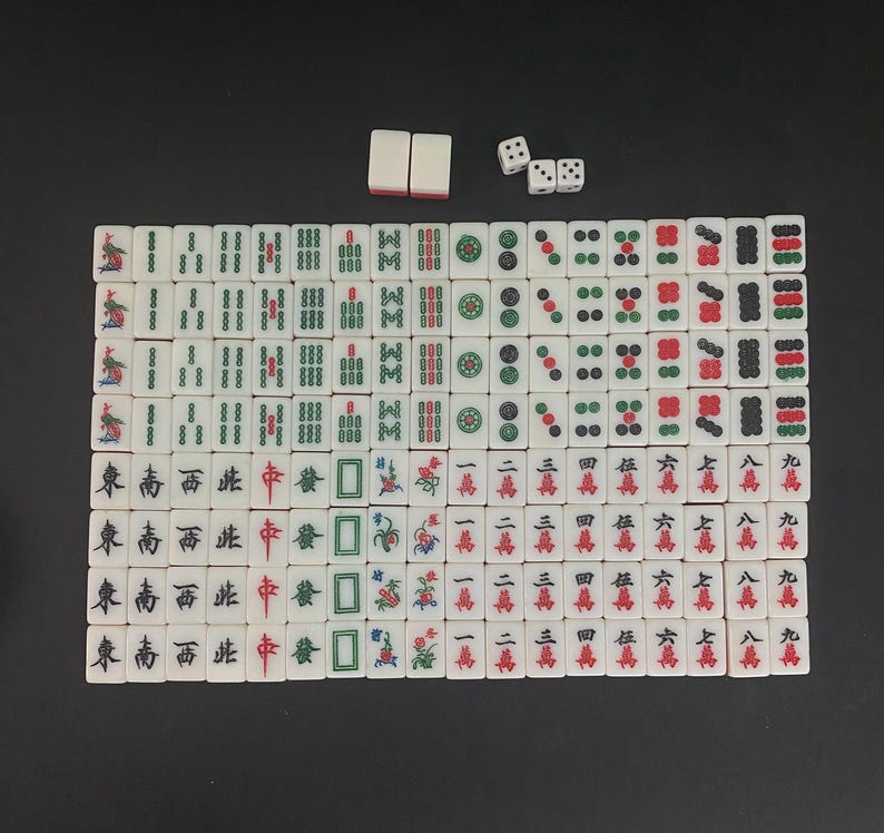 Puede incluir: Un juego de Mahjong completo con fichas blancas que presentan varios s&iacute;mbolos y caracteres en rojo, verde y negro. Incluye cuatro dados y dos fichas de repuesto. Las fichas est&aacute;n dispuestas sobre una superficie oscura.