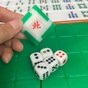 May include: A close-up of a Mahjong game in progress. A green and white tile with a red symbol is held above a set of white dice with black dots and one red-dotted die. The game tiles are green and white.
