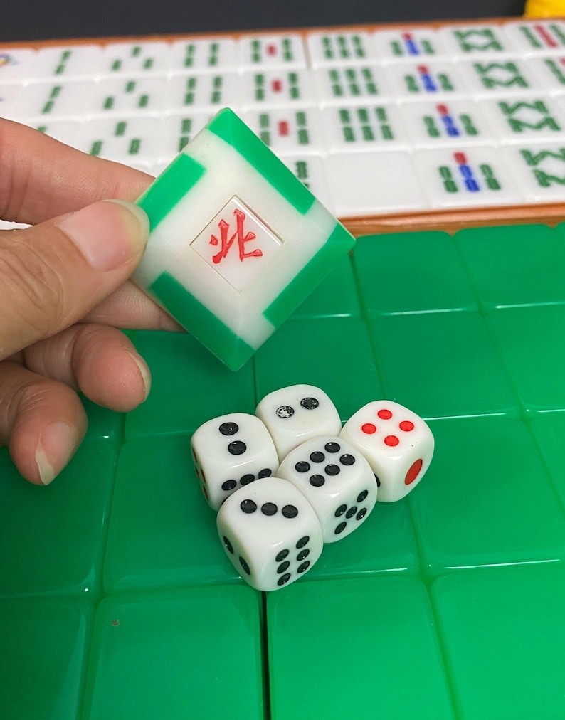 May include: A close-up of a Mahjong game in progress. A green and white tile with a red symbol is held above a set of white dice with black dots and one red-dotted die. The game tiles are green and white.