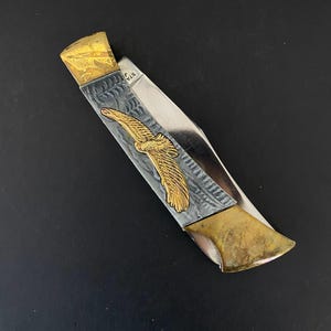 May include: A folding pocket knife with a gold eagle emblem on a silver and gray handle. The knife has a gold-colored blade end and a stainless steel blade. The knife is on a black surface.