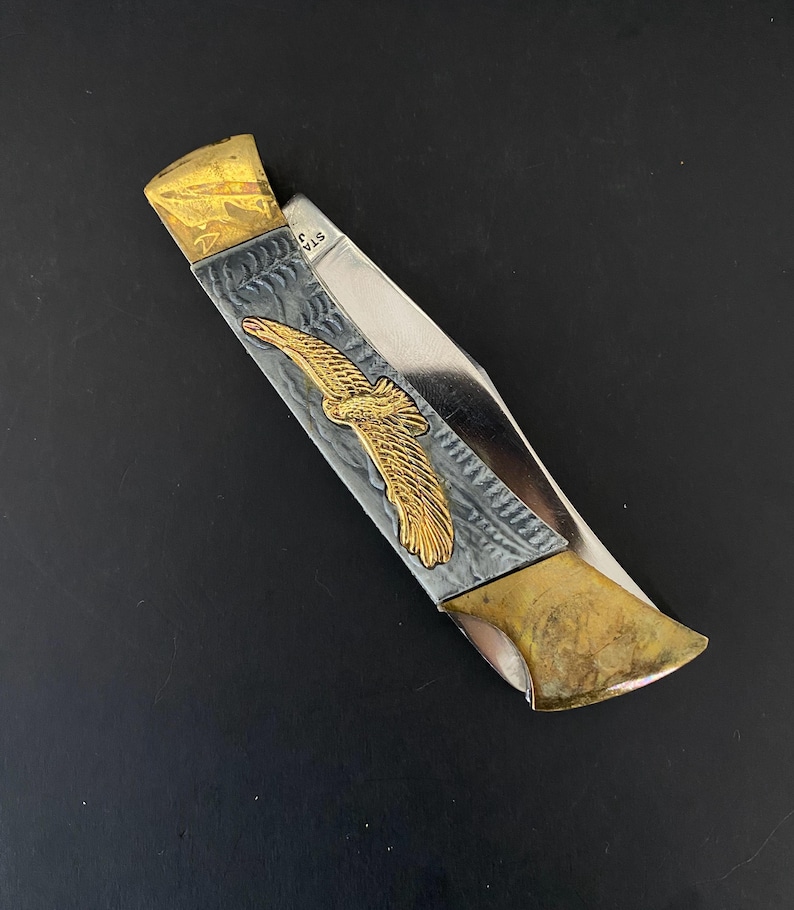 May include: A folding pocket knife with a gold eagle emblem on a silver and gray handle. The knife has a gold-colored blade end and a stainless steel blade. The knife is on a black surface.