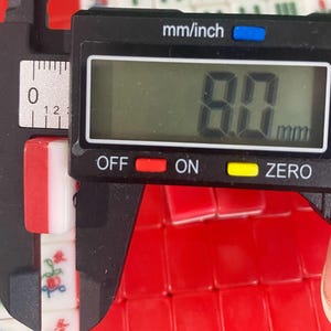 Puede incluir: Una ficha roja y blanca de un juego de Mahjong se mide con un calibrador digital. La ficha mide 8,0 mm de ancho. El calibrador muestra la medida en mil&iacute;metros. El fondo est&aacute; lleno de fichas rojas.