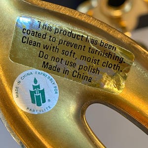 May include: Close-up of a gold-colored metal decorative object with a sticker that reads "This product has been coated to prevent tarnishing. Clean with soft, moist cloth. Do not use polish. Made in China."