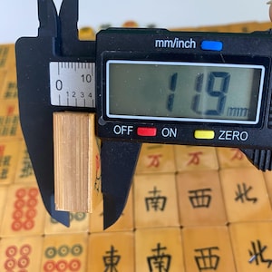 May include: A digital caliper measures the thickness of a small, rectangular, light-colored object. The caliper displays a measurement of 11.9 mm. The object is surrounded by other similar objects with Chinese characters.