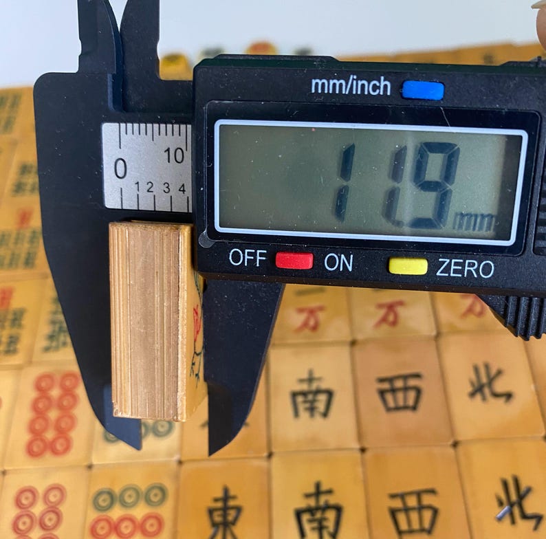 May include: A digital caliper measures the thickness of a small, rectangular, light-colored object. The caliper displays a measurement of 11.9 mm. The object is surrounded by other similar objects with Chinese characters.