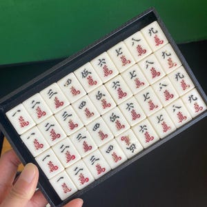 May include: A set of white Mahjong tiles with red and black markings, neatly arranged in a black tray. The tiles display Chinese characters and numbers, perfect for game enthusiasts.