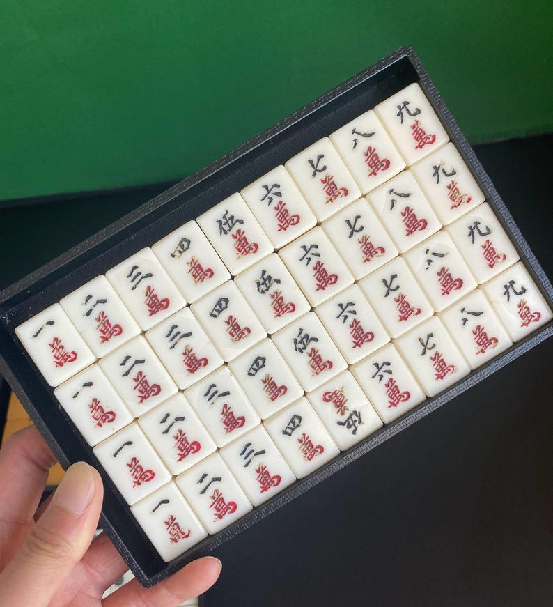 May include: A set of white Mahjong tiles with red and black markings, neatly arranged in a black tray. The tiles display Chinese characters and numbers, perfect for game enthusiasts.