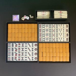 May include: A complete Mahjong set with tiles, dice, and a wind indicator. The tiles are arranged in black trays, with a mix of ivory and orange tiles. The dice are white with black dots. The wind indicator is a purple square.