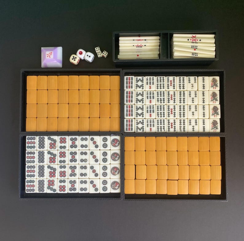 May include: A complete Mahjong set with tiles, dice, and a wind indicator. The tiles are arranged in black trays, with a mix of ivory and orange tiles. The dice are white with black dots. The wind indicator is a purple square.