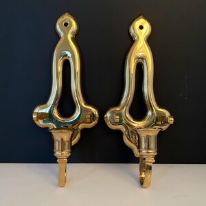 May include: Two ornate, polished brass wall sconces. Each sconce features a decorative top with a central opening, a flared midsection, and a candle cup with a hook. The sconces are displayed against a dark background.