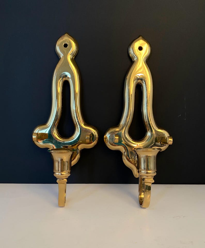 May include: Two ornate, polished brass wall sconces. Each sconce features a decorative top with a central opening, a flared midsection, and a candle cup with a hook. The sconces are displayed against a dark background.