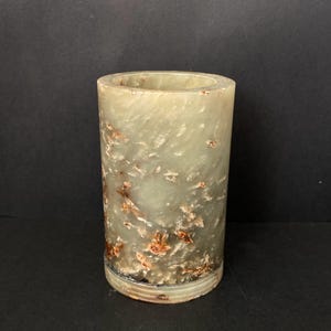 Könnte beinhalten: Eine zylindrische grüne Jadevase mit braunen und weißen Markierungen. Die Vase hat eine glatte, polierte Oberfläche.