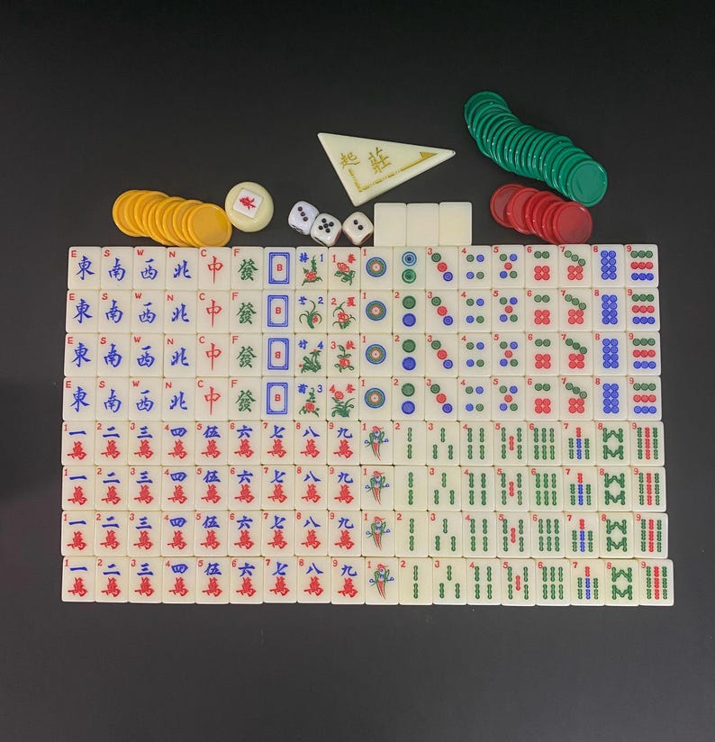 May include: A complete Mahjong set with ivory-coloured tiles, dice, and coloured chips. The tiles feature Chinese characters and symbols in red, green, and blue. The set includes green, red, and yellow chips, and a white triangular piece.