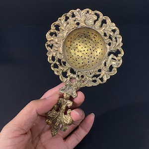 May include: A brass tea strainer featuring a circular bowl with a perforated base and an intricate, ornate filigree border. The handle is shaped with detailed scrollwork and decorative metal patterns, showcasing a metallic gold finish.