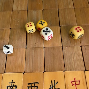 May include: A close-up shot of a wooden surface with several dice and Mahjong tiles. The dice are yellow and white with black and red dots. The Mahjong tiles are brown with Chinese characters.