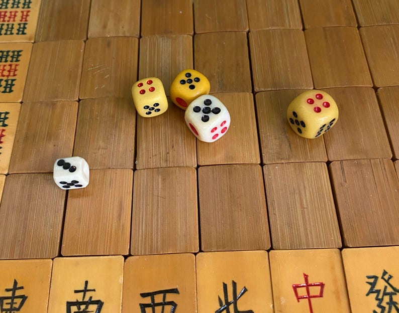 May include: A close-up shot of a wooden surface with several dice and Mahjong tiles. The dice are yellow and white with black and red dots. The Mahjong tiles are brown with Chinese characters.
