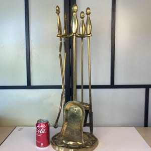 May include: A vintage brass fireplace tool set. The set includes a shovel, poker, brush, and tongs, all with decorative handles. The tools are displayed on a matching base. A red soda can is next to the set for size comparison.