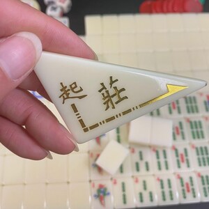 May include: A cream-coloured Mahjong tile with gold Chinese characters and a gold arrow design. The tile is triangular and is held in a hand, with other Mahjong tiles visible in the background.
