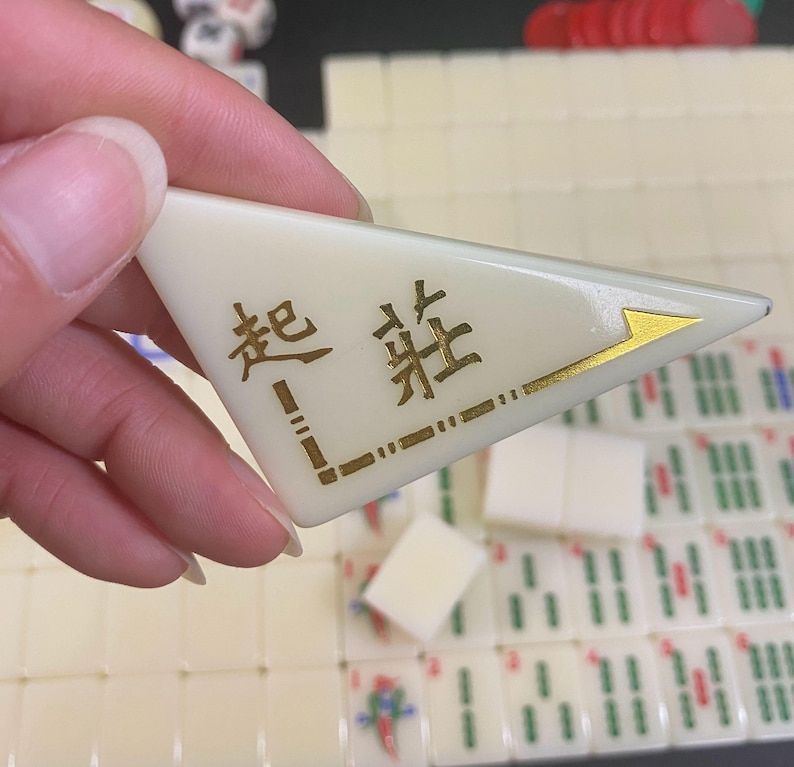 May include: A cream-coloured Mahjong tile with gold Chinese characters and a gold arrow design. The tile is triangular and is held in a hand, with other Mahjong tiles visible in the background.