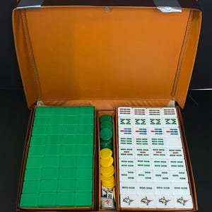 May include: A vintage Mahjong set in an open brown case. The set contains green tiles, yellow and green chips, dice, and white tiles with red, green, and blue markings. The case is open, displaying the game pieces.