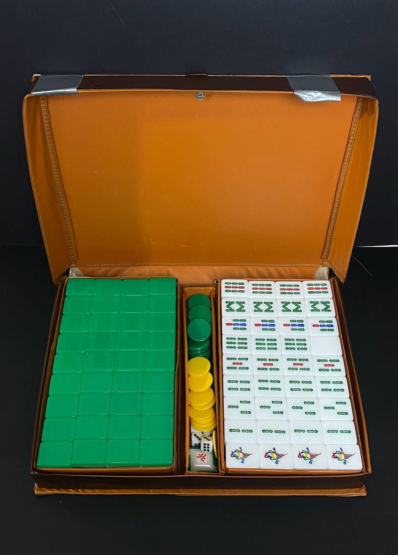 May include: A vintage Mahjong set in an open brown case. The set contains green tiles, yellow and green chips, dice, and white tiles with red, green, and blue markings. The case is open, displaying the game pieces.