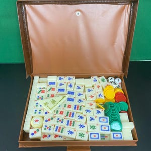 May include: A Mahjong set in a brown case, open to reveal the game pieces. The ivory-coloured tiles have red, green, blue, and black symbols and characters. The set also includes dice and coloured chips. The case is made of leatherette.