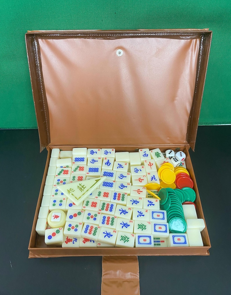 May include: A Mahjong set in a brown case, open to reveal the game pieces. The ivory-coloured tiles have red, green, blue, and black symbols and characters. The set also includes dice and coloured chips. The case is made of leatherette.