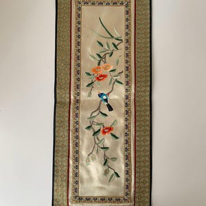 May include: Embroidered wall hanging featuring a bird perched on a flowering branch. The artwork has a cream-colored background with orange and green floral details. The border is green with gold accents and a black trim.