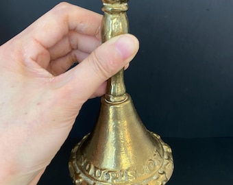 May include: A vintage brass bell with a flared base and a long handle. The bell has an embossed design around the base and a small knob at the top. The bell is held in a hand against a dark background.