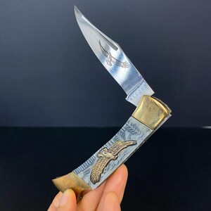 May include: A silver and gold folding knife with an eagle design. The blade is partially open, revealing an etched eagle. The handle features an embossed eagle and decorative patterns. The text "KEE098" is visible on the blade.