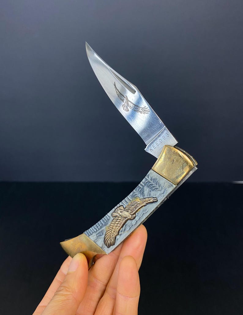 May include: A silver and gold folding knife with an eagle design. The blade is partially open, revealing an etched eagle. The handle features an embossed eagle and decorative patterns. The text "KEE098" is visible on the blade.