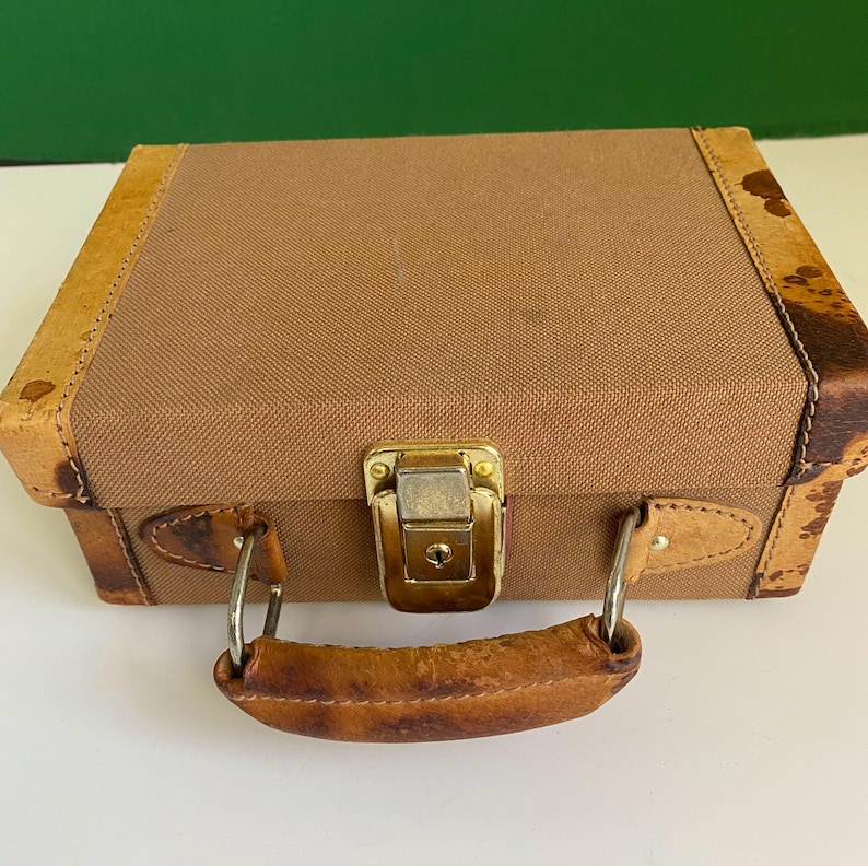 May include: A small, vintage brown suitcase with a canvas body and leather trim. The suitcase has a gold-colored metal clasp and a leather handle. The corners of the suitcase are reinforced with patterned leather. The handle is attached with silver-colored metal hardware.