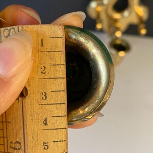 May include: Close-up of a brass-colored object with a green interior, being measured with a wooden ruler. The ruler shows measurements in inches. The object appears to be approximately 2 inches in diameter.