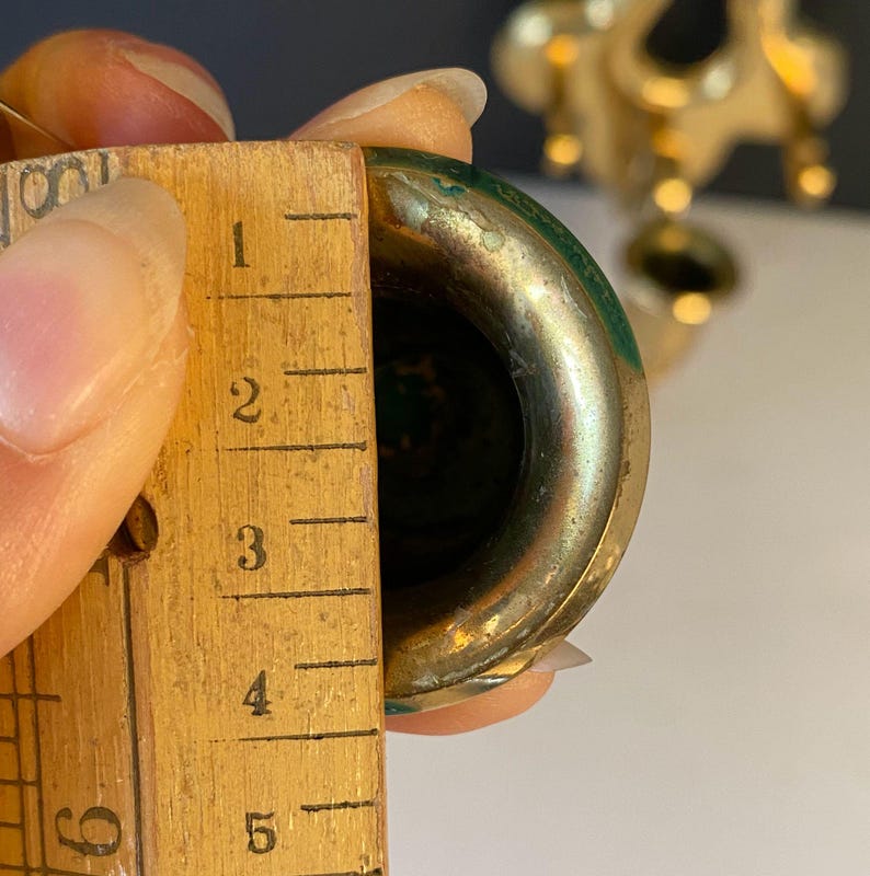 May include: Close-up of a brass-colored object with a green interior, being measured with a wooden ruler. The ruler shows measurements in inches. The object appears to be approximately 2 inches in diameter.